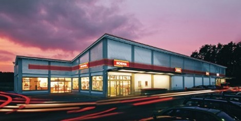 Das Bild zeigt ein modernes Supermarktgebäude bei Abenddämmerung. Das Gebäude ist hell beleuchtet und trägt das auffällige rote NORMA-Logo über den Eingängen. Im Vordergrund sind Lichtspuren vorbeifahrender Autos zu sehen, was dem Bild eine dynamische Wirkung verleiht. Der Himmel ist in violett-rosa Tönen gefärbt, was eine stimmungsvolle Atmosphäre erzeugt.