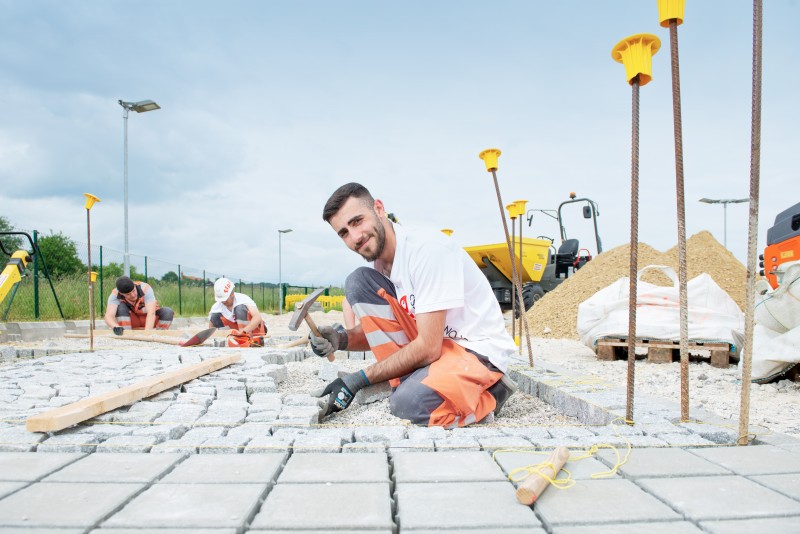 Das Bild zeigt einen jungen Mann bei Pflasterarbeiten im Freien. Er kniet auf dem Boden und verlegt rechteckige Betonsteine. Im Hintergrund sind weitere Arbeiter, Baumaterialien und Werkzeuge zu sehen. Die Szene wirkt professionell und zeigt einen typischen Arbeitstag auf einer Baustelle.